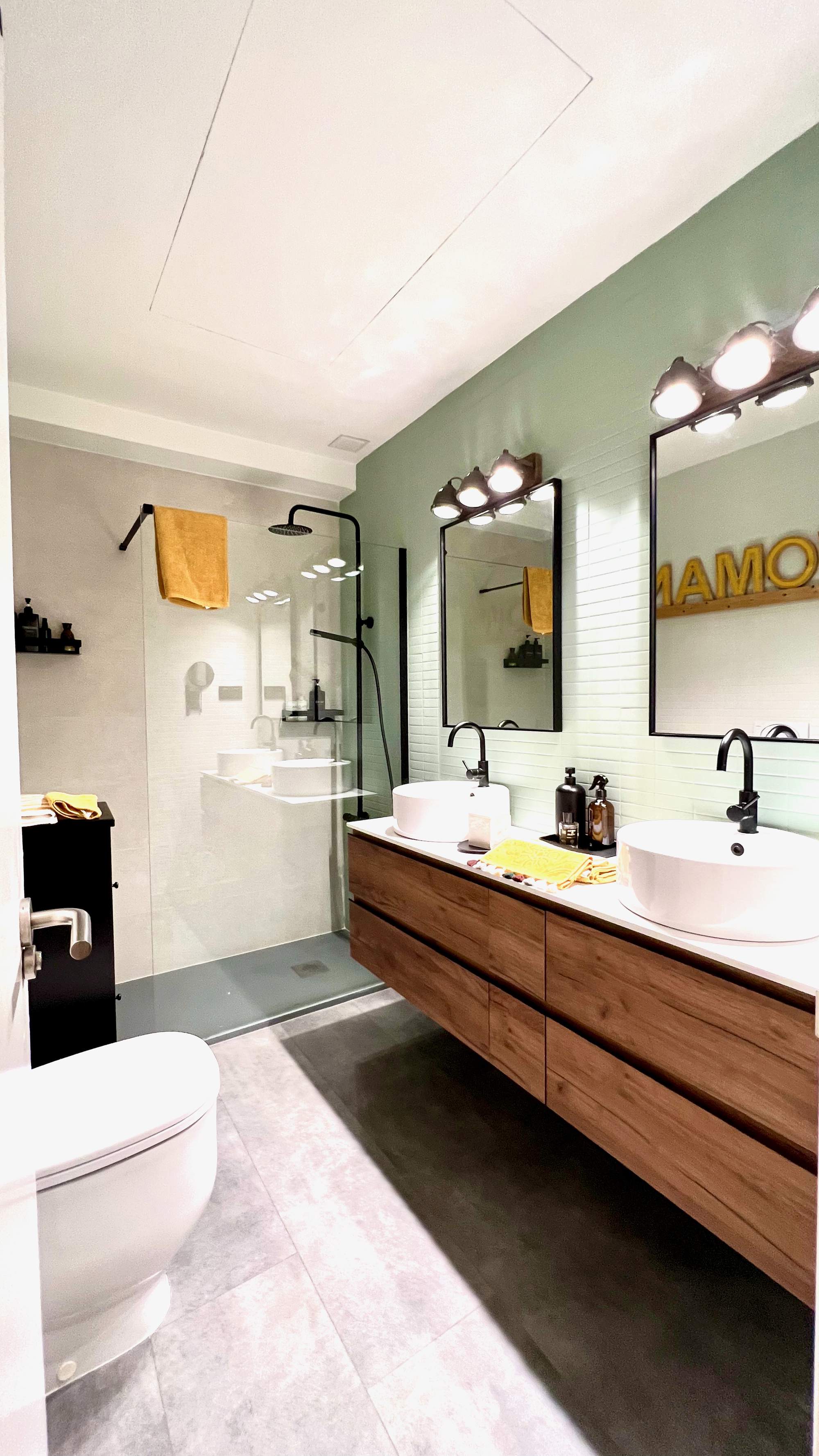 Baño con espejos iluminados, mueble con doble lavabo de madera y ducha al fondo.