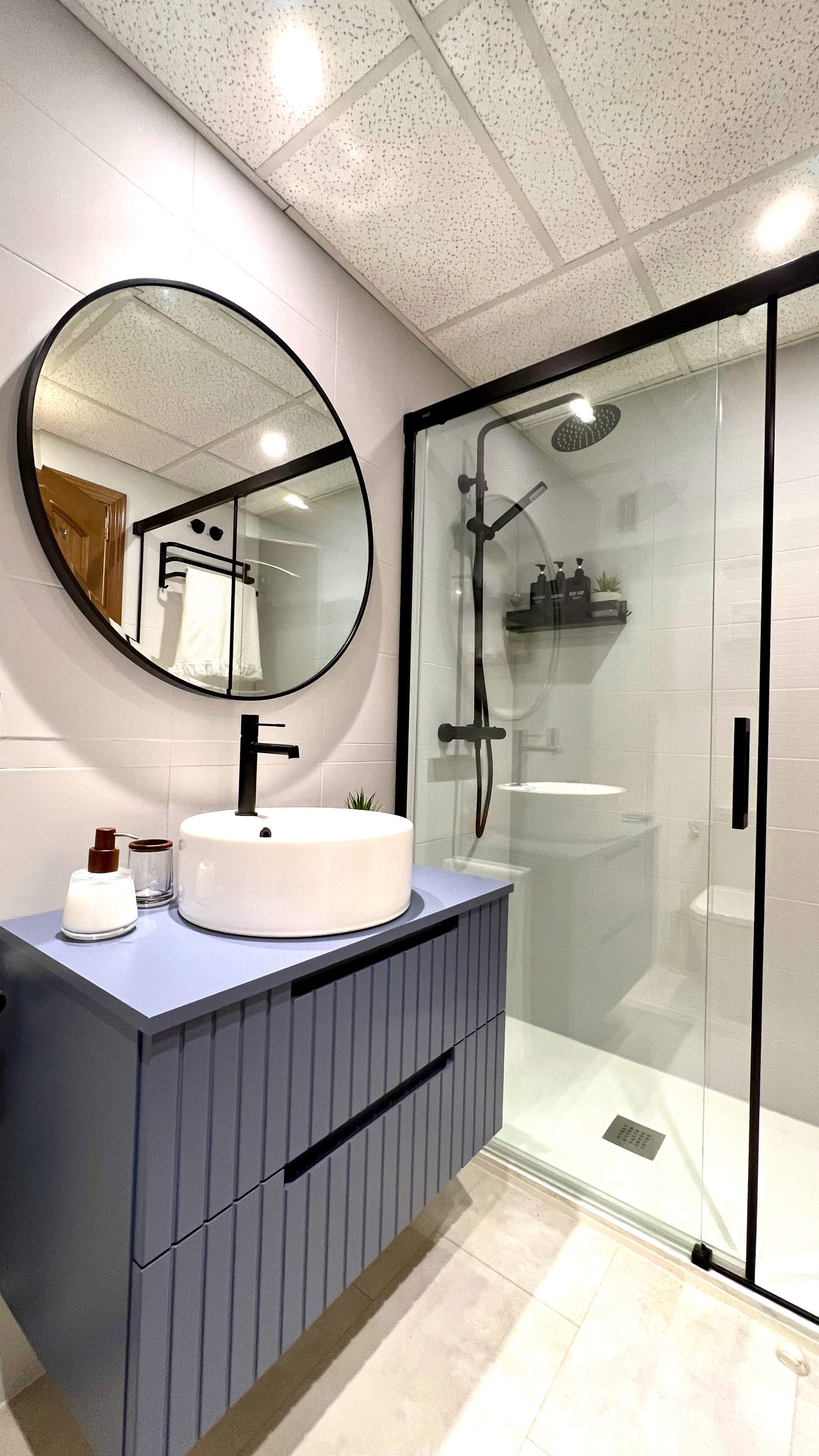 Baño con espejo redondo, mueble bajolavabo azul de madera y ducha con mampara.