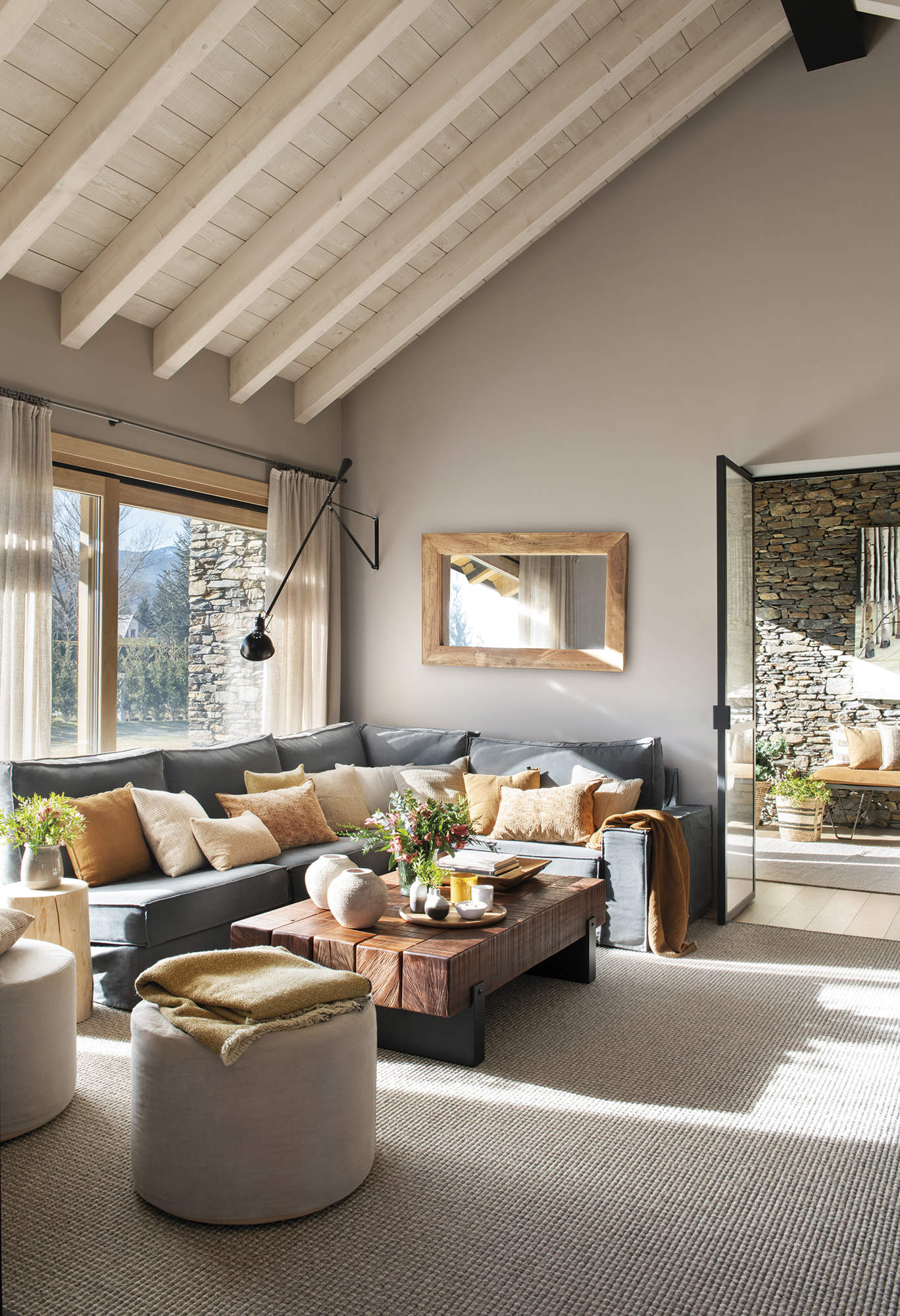 Salón con techo alto con vigas de madera, sofá gris esquinero con cojines beige y naranja, mesa de centro de madera rústica,  alfombra gris.