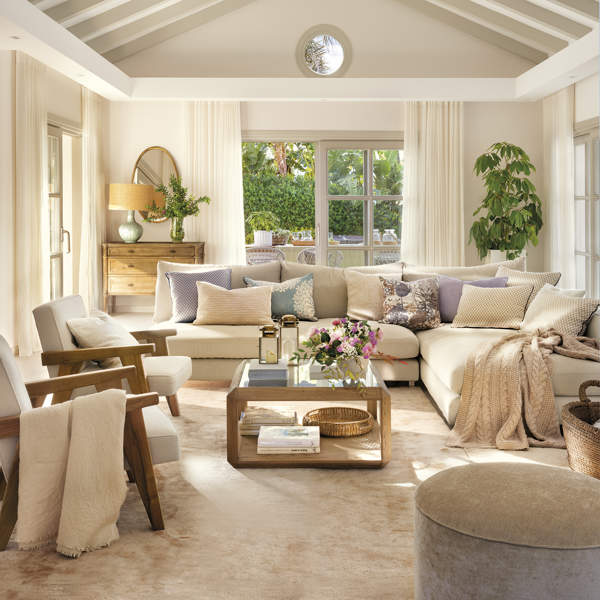 salón blanco con techo a dos aguas con vigas de madera beige, sofá esquinero, butacas de madera tapizadas, alfombra beige, puf